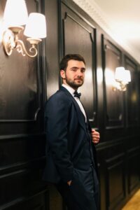 A stylish man in a tuxedo leans against an elegant hallway wall under warm lighting.